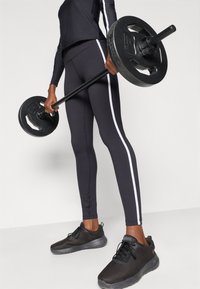 Top a maniche lunghe nera e leggings con strisce bianche sui lati, che tiene un bilanciere con pesi neri. Scarpe da ginnastica nere in mesh completano l'outfit.
