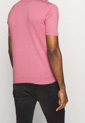 Mann, der ein eng anliegendes rosa Kurzarmhemd und schwarze Jeans trägt, von hinten vor schlichtem Hintergrund gezeigt.