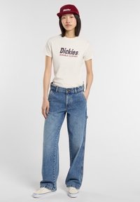 Witte T-shirt met "Dickies" en "Duurzame Kleding" in donkerbruine tekst, blauwe wijde jeans en bordeauxrode corduroy pet. Crème sneakers.