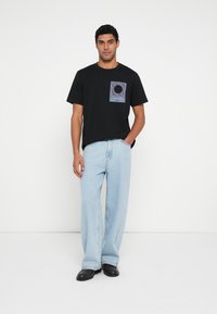 Melna T-krekls ar grafisku izdruku un vertikālām svītrām, kopā ar gaiši zilām platām biksēm un melniem apaviem. Vienkāršs dizains, ikdienišķs stils.