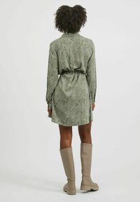 Kvinna med lockigt hår iklädd en grönmönstrad långärmad klänning och beige knähöga stövlar, stående vänd bortåt mot en neutral bakgrund.