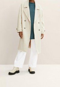 Manteau beige à chevrons avec boutons croisés, associé à un pull en maille bleu sarcelle, un pantalon large blanc et des bottes à plateforme crème.