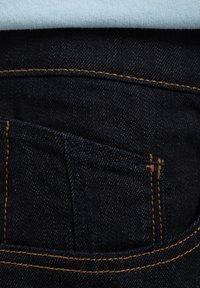 Ciemne dżinsy z przodu z detalem kieszeni, ozdobione złotym przeszyciem na szwach. Powyżej częściowo widoczna jasnoniebieska tkanina.