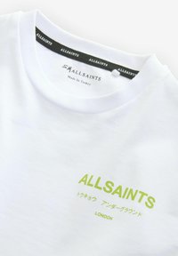 Balts kokvilnas T-krekls ar apaļu kaklu, zaļu "ALLSAINTS" logo un japāņu teksta dizainu. Etiķete norāda "Ražots Turcijā."