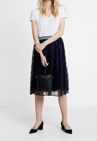 Granatowa koronkowa spódnica midi nałożona na gładką białą koszulkę z dekoltem w serek, w połączeniu z czarnymi butami na słupku i małą czarną torebką.