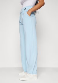 Lichtblauwe wijde broek met een hoge taille, voorzien van zijzakken, plooien en een zwart knoopdetail. G gedragen met witte sneakers.