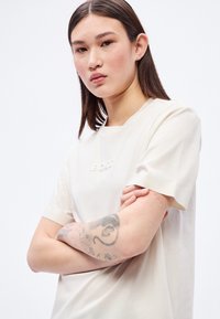 Κρεμ κοντομάνικη μπλούζα με ανάγλυφο λογότυπο "BOSS". Το ύφασμα φαίνεται μαλακό, με άνετη εφαρμογή και μινιμαλιστικό σχεδιασμό.