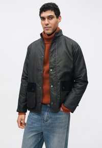 Fekete steppelt dzseki magas gallérral, patentos gombokkal, két elülső zsebbel és kontrasztos sötét szövetes részletekkel. Narancssárga garbó felett viselve.