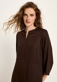 Frau mit welligem roten Haar, die ein dunkelbraunes Langarmkleid mit einem Reißverschlussdetail vorne trägt, steht vor einem schlichten beigen Hintergrund.