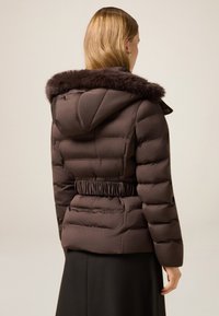 Brązowa pikowana kurtka z sztucznym futrzanym kapturem, elastycznym paskiem w talii i poziomym pikowaniem. Charakteryzuje się gładką fakturą i uporządkowanym krojem.