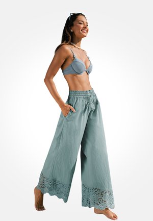 Femme souriante, marchant pieds nus, portant un haut de soutien-gorge bleu clair et un pantalon ample à jambes larges avec bordure en dentelle aux ourlets et les mains dans les poches.