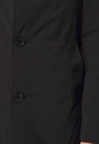 Zbliżenie czarnego płaszcza pokazujące dwa czarne guziki i boczną kieszeń po prawej stronie, noszony przez osobę, której widoczne są nadgarstek i ręka.