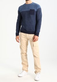 Pull en tricot dans des tons bleus avec un motif texturé et une poche avant, associé à un pantalon cargo beige et des baskets blanches.