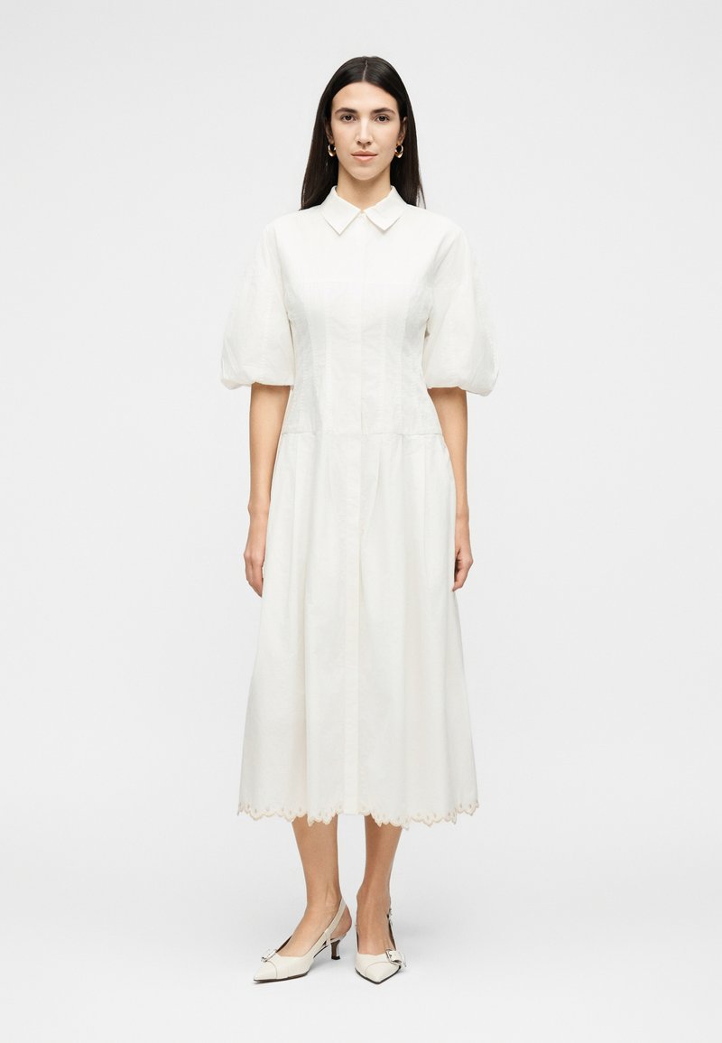 Γυναίκα που στέκεται, φοράει άσπρο μίντι φόρεμα με φουσκωτά μανίκια, μυτερό γιακά, κυματιστό τελείωμα και άσπρα πέδιλα slingback με φιόγκους.