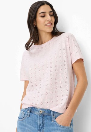 Junge Frau trägt ein hellrosa Ösen-Top mit kurzen Ärmeln und blaue Jeans, die Hände in den Taschen, schaut nach unten und lächelt leicht.