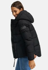 Black puffer jacket with a large hood, zip closure, and a logo patch on the sleeve. Smooth texture and quilted design with a fitted silhouette.