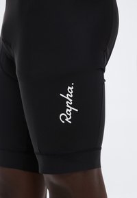Μαύρο ποδηλατικό σορτς Rapha φορεμένο στον μηρό, με λευκό λογότυπο "Rapha" στο πλάι πάνω σε ουδέτερο φόντο.