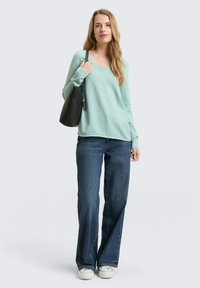 Hellgrüner, langärmliger Pullover mit V-Ausschnitt, kombiniert mit dunkelblauen, weit geschnittenen Jeans und weißen Turnschuhen. In einer Hand wird eine schwarze Tragetasche gehalten.