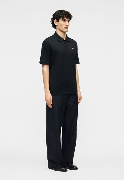 Άνδρας που στέκεται φορώντας μαύρο πόλο μπλουζάκι με κοντό μανίκι, μαύρο παντελόνι με ρίγες και φαρδιά μπατζάκια, και μαύρα παπούτσια, μπροστά σε ένα απλό φόντο.