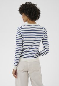 Femme aux cheveux bouclés portant un pull à manches longues blanc à rayures bleues et un pantalon beige clair, debout de dos face à la caméra.