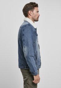 Giacca in denim di blu medio con colletto in ecopelliccia bianca, dotata di tasche laterali, chiusura a bottoni e una finitura leggermente usurata.