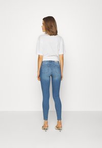Jeans skinny azuis com uma textura suave, apresentando bolsos traseiros. Combinados com uma t-shirt branca de mangas curtas e sandálias de salto.