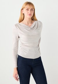 Długi rękaw, top z kapturem w jasnobeżowym, teksturowanym materiale z subtelnym połyskiem. Podkreślony marszczeniem w talii.
