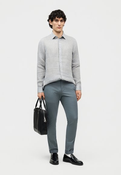 Νέος με σγουρά μαλλιά που φοράει ανοιχτό γκρίζο πουκάμισο με κουμπιά, μπλε-γκρίζο παντελόνι, μαύρα μοκασίνια και κρατά μια μαύρη τσάντα εργασίας.