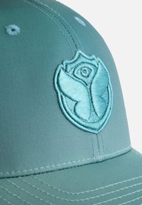 Teal pet met een geborduurd embleem met een bloemenontwerp. De stof heeft een gladde textuur en zichtbare stiksels rond de randen.