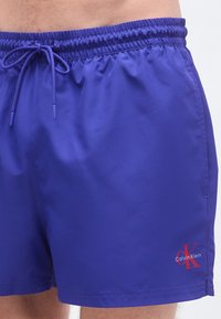 Ανδρικό μαγιό σορτς σε ζωντανό μοβ ύφασμα. Διαθέτει ελαστική μέση με κορδόνι, τσέπες στα πλαϊνά ραφές και κόκκινο λογότυπο ως διακριτικό.