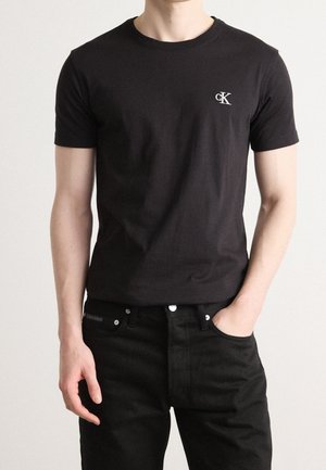 Mężczyzna w dopasowanej czarnej koszulce Calvin Klein i czarnych dżinsach, stojący z jedną ręką w kieszeni na jednolitym tle.
