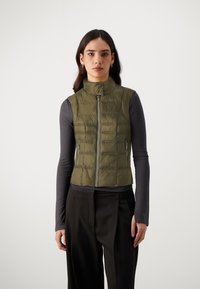 Gilet imbottito verde oliva con colletto alto, chiusura zip davanti e tasche laterali con zip. Indossato sopra un maglione scuro a maniche lunghe, con una texture liscia.