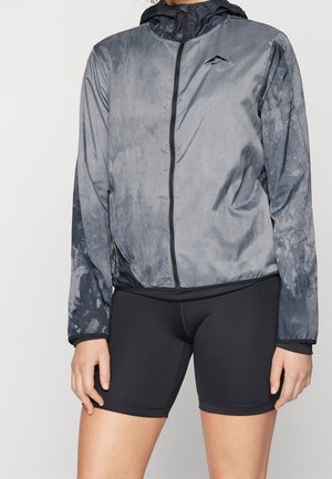 Person trägt eine grau-schwarze Jacke mit Reißverschluss und schwarze enganliegende Shorts und steht vor einem einfarbigen Hintergrund.