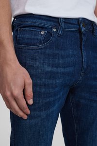 Temno modri jeans iz denima z gladko teksturo, z dvema sprednjima žepoma, kovinsko gumbom in diskretnim detajlom zasekanja. Podrobni pogled na prileganje.