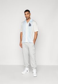 Kurzarmhemd in Hellblau und Beige mit einem marineblauen "LA"-Logo. Kombiniert mit grauen Cargo-Sweatpants und weißen Sneakers.