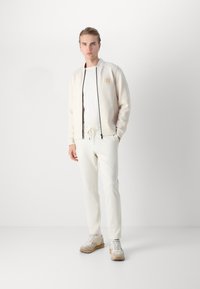 Light-colored zip-up bomber jacket with a round logo, paired with matching pants and beige sneakers. Smooth fabric, simple design.