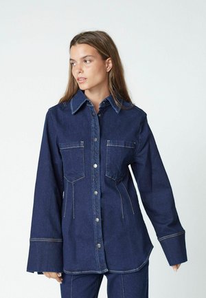 Denim button-up shirt, dark blue with two chest pockets, a pointed collar, and contrast stitching, featuring wide sleeves and a curved hem.