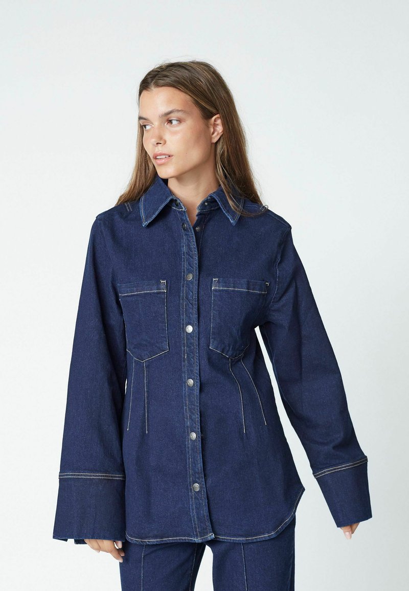 Denim button-up shirt, dark blue with two chest pockets, a pointed collar, and contrast stitching, featuring wide sleeves and a curved hem.