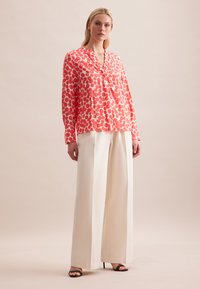 Blusa a pois rossi e bianchi con scollatura a V, abbinata a pantaloni beige a gamba larga con vita alta e tacchi aperti neri.