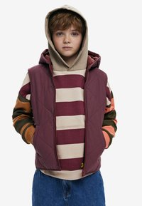 Vadderad maroon ärmlös jacka över en randig beige och burgundy hoodie. Hoodien har orange och gröna accenter på ärmarna. Blå jeans.