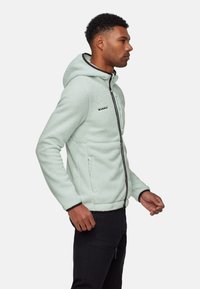 Mann, der einen hellgrünen Fleece-Reißverschlusskapuzenpullover mit hochgestellter Kapuze trägt, im Seitenprofil vor weißem Hintergrund steht.