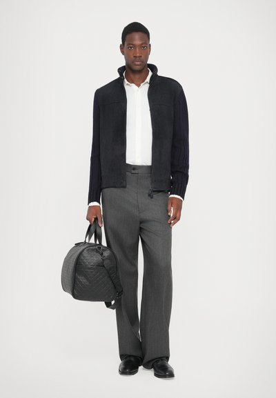 Veste en daim noire, chemise blanche à boutons, pantalon large gris, et un sac de voyage noir texturé avec double poignées. Chaussures noires lisses.