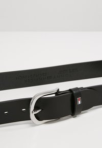 Ceinture en cuir noir avec une boucle en métal argenté. Présente un logo sur le passant, avec des trous pour l'ajustement de taille, texture lisse.