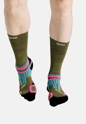 Benen met olijfgroene compressiekousen met roze, blauwe en zwarte accenten, die de achterkant van de kuiten en voeten tonen op een witte achtergrond.