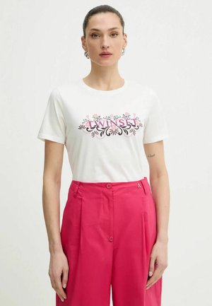 Donna con capelli scuri raccolti in uno chignon che indossa una maglietta bianca con la scritta "SUNSET" e un motivo floreale, abbinata a pantaloni a vita alta di colore rosa acceso.