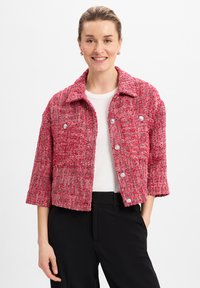 Rote und pinke Tweedjacke mit verkürztem Design, Kragen, Vorderknöpfen und zwei Brusttaschen; getragen über einem weißen Hemd und schwarzen Hosen.