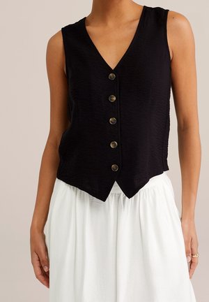 Gilet noir sans manches à boutons avec col en V, porté sur une jupe blanche froncée, visible des épaules jusqu'aux hanches.