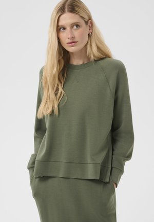 Femme blonde portant un sweat à manches longues vert olive et un bas assorti, les mains dans les poches, regardant légèrement sur le côté.