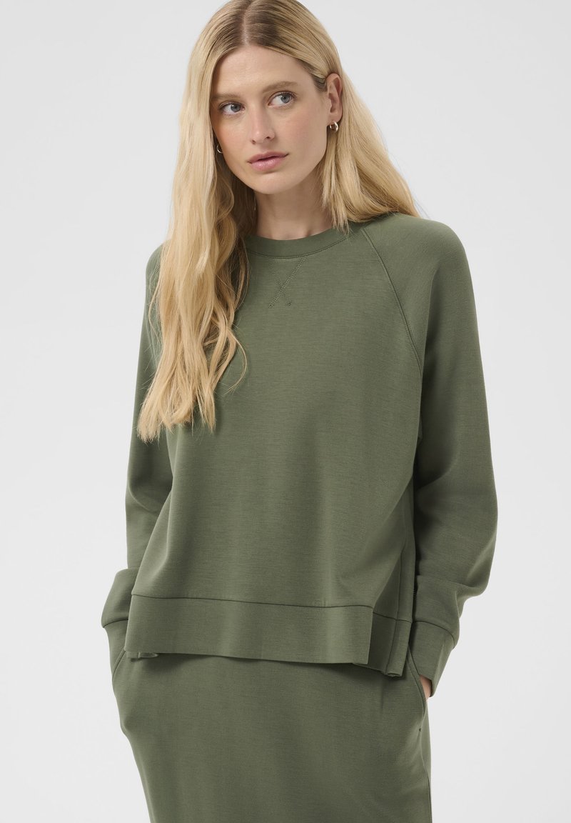 Femme blonde portant un sweat à manches longues vert olive et un bas assorti, les mains dans les poches, regardant légèrement sur le côté.