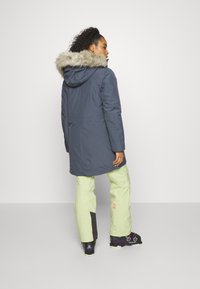 Námořnická parka s kožešinovou podšívkou, dlouhého střihu, v kombinaci se světle zelenými kalhotami, které mají černý akcent a černou obuv s růžovými detaily.